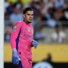Bramkarz Manchester City w różowym stroju, z niebieskimi rękawicami, gotowy do obrony bramki.