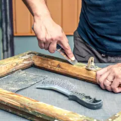 Mani esperte usano uno raschietto per sverniciare legno, rimuovendo vecchie vernici da un telaio. Accanto, una spazzola metallica e carta vetrata.