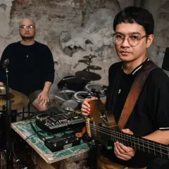 Dwóch muzyków w surowym wnętrzu, jeden gra na gitarze, drugi siedzi przy perkusji. Brzmienie tutti jest gotowe do nagrania.