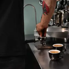 Barista z tatuażem na ramieniu przygotowuje kawę americana. Dwie filiżanki czarnej kawy stoją na blacie obok ekspresu.