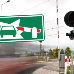 Zielony znak informuje o oznaczeniu przejazdu kolejowego. Po bokach widać rogatki i sygnalizację świetlną.
