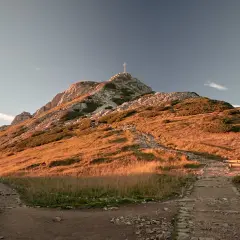 Kamienista trasa na Giewont w złotym świetle zachodzącego słońca. Widoczny krzyż na szczycie i ławki dla turystów.