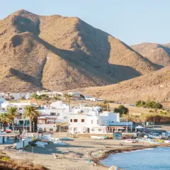 Vista de la finca Maltes del Fraile, con casas blancas, palmeras y montañas áridas al fondo. Barcas en la playa y el mar azul.