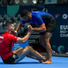 Två män tävlar i ju jitsu. En sitter på mattan i röd dräkt, den andra står i blå dräkt och sträcker ut en hand.