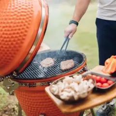 Un homme cuisine des steaks sur un kamado, c'est quoi ce barbecue en céramique orange. Des légumes sont prêts à être grillés.