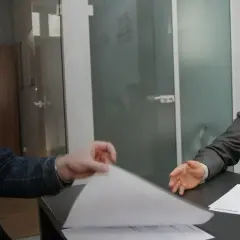 Dwóch mężczyzn przy biurku przegląda dokumenty, jeden w garniturze, drugi w dżinsowej kurtce.