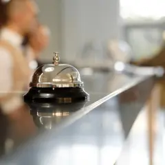 Dzwonek hotelowy na ladzie, symbol usług uzupełniających. W tle niewyraźne postacie personelu.