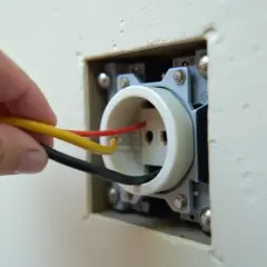 Dłoń z przewodami (żółty, czerwony, czarny) zbliża się do gniazdka elektrycznego.