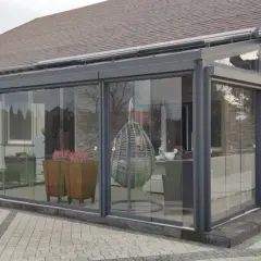 Nowoczesny taras zimowy z przeszklonymi ścianami, wiszącym fotelem i donicami z wrzosami. Idealne miejsce na relaks o każdej porze roku.