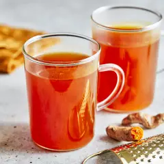 Zwei Gläser mit leuchtend orangefarbenem Getränk, das nach einem Kurkuma Honig Rezept zubereitet wurde. Frische Kurkumawurzeln und ein Sieb liegen daneben.