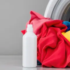 Bouteille de vinaigre blanc à côté d'une machine à laver remplie de linge coloré. Idéal pour enlever la colle tenace des textiles.