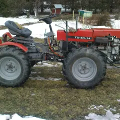 Czerwony traktor TZ-4K-14 z dużymi kołami stoi na trawiastym podwórku, częściowo pokrytym śniegiem.