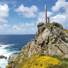 Escaleras de piedra ascienden por rocas cubiertas de flores amarillas hasta un monumento en el cabo Priorino. El mar azul y el cielo con nubes completan la escena.