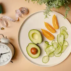 Zegar, taśma miernicza i talerz z awokado, pomidorem i ogórkiem symbolizują dietę imitującą post i zdrowy styl życia.