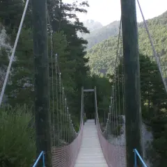 Puente colgante de madera y metal en la vía del Nicolau, rodeado de bosques y montañas.