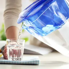 Une personne verse de l'eau filtrée d'une carafe bleue dans un verre. À côté, un citron coupé. L'eau filtrée est elle bonne pour la santé ?