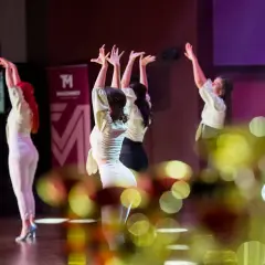 Tancerki w jasnych bluzkach i białych spodniach wykonują zmysłowy taniec. W tle logo szkoły tańca. To piękna **bachata solo**.