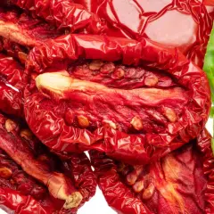 Tomates secos al horno, arrugados y rojos, con semillas visibles, junto a una hoja de albahaca verde fresca.