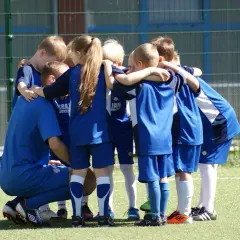 Dzieci w niebieskich strojach piłkarskich tworzą kółko. Wśród nich klęczy asystent trenera, wspierając młodych zawodników.