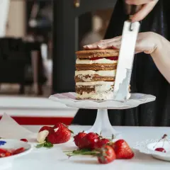 Osoba dekoruje tort, nakładając na niego prosty tynk do tortu. Na stole leżą świeże truskawki i wiśnie.