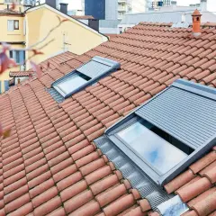 Tegole rosse su un tetto con una pendenza tetto 30 gradi, due lucernari con tapparelle grigie parzialmente abbassate.