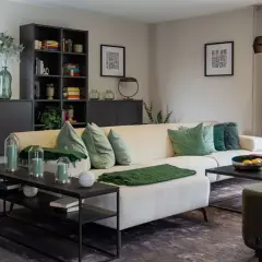 Gemütliches Wohnzimmer mit cremefarbener Couch, grünen Kissen und Decken. Helle Farben dominieren, ergänzt durch dunkle Möbel und eine große Glasfront.