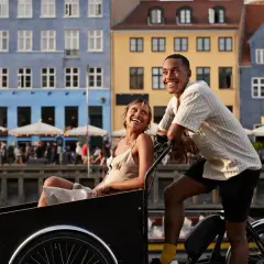 Ett lyckligt par njuter av en romantisk weekend i Europa, cyklar längs en kanal med färgglada hus i bakgrunden.