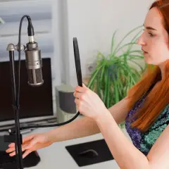 Kobieta z rudymi włosami i słuchawkami ustawia mikrofon studyjny. Dowiedz się, jak podłączyć mikrofon do laptopa, aby nagrywać dźwięk.