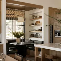 A cozy breakfast nook built in with a dark table, plush seating, and a large window. Shelves display rustic pottery and kitchenware.