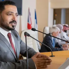 A man speaks at a podium, perhaps sharing insights from his pursuit of best online MPA programs, as others listen at a long table with flags.