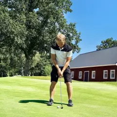 Man puttar på en golfbana, en röd byggnad och träd i bakgrunden. Golf som friskvård är en härlig aktivitet.