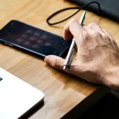 Dłoń trzyma długopis nad ekranem z klawiaturą numeryczną, gdzie urządzenie iPhone niedostępne. Obok leży laptop.