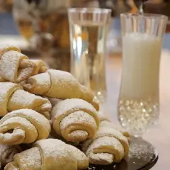 Stos puszystych rogalików krucho drożdżowych, posypanych cukrem pudrem, obok kieliszków z napojem.