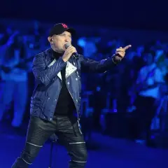 Charyzmatyczny de mono wokalista w czapce z daszkiem i skórzanych spodniach śpiewa na koncercie, wskazując palcem na publiczność.