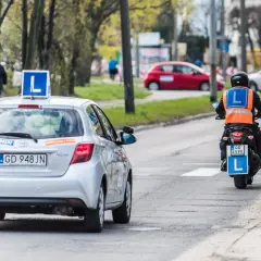 Srebrny samochód nauki jazdy z literą "L" na dachu i motocyklista w kamizelce odblaskowej.