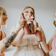 Trzy uśmiechnięte kobiety w różowych sukienkach wznoszą toasty błyszczącymi butelkami. Radosne przygotowania do ślubu.