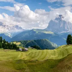 Zielone wzgórza i drewniane chatki w Val Gardena, z majestatycznymi Dolomitami w tle.