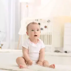 Małe dziecko z niebieskimi oczami siedzi na białym dywanie, odkrywając świat. To piękny moment, kiedy dziecko zaczyna siadać.