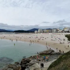 Gente disfruta de un día soleado en la playa de peizas, con aguas cristalinas y edificios al fondo.