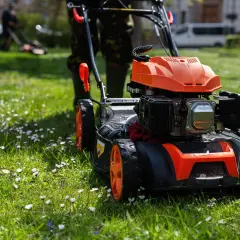 Kosiarka polskiego producenta kosiarek z pomarańczowymi detalami na tle zielonej trawy z kwiatami.