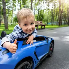Uśmiechnięty chłopiec daje kciuk w górę, siedząc w niebieskim samochodzie dla dziecka zabawce na tle parku.