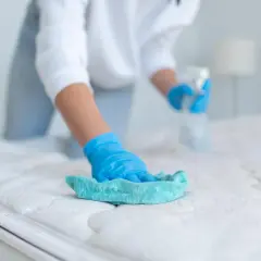 Persona con guantes azules limpia un colchón con un paño turquesa y spray. Aprende como limpiar colchones en casa para un descanso higiénico.