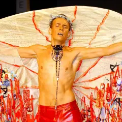 Mężczyzna w koronie cierniowej i czerwonych spodniach, z rozpostartymi ramionami, prezentuje ekstrawagancki strój. To dzieło Arkadiusza projektanta, pełne symboli.