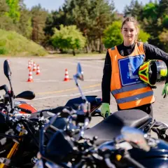 Młoda kobieta w kamizelce odblaskowej z literą L trzyma kask na placu manewrowym z motocyklami.