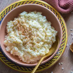 Omas Milchreis mit Butter, bestreut mit Zimt, in einer Schüssel auf einem Holztisch. Daneben eine kleine Schale Reis und eine Zimtstange.