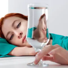Mujer pelirroja recostada, con su rostro reflejado en un vaso de agua, quizás pensando en los beneficios de un alcalinizador de agua para su bienestar.