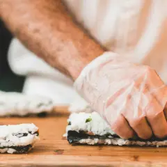 Hände mit Handschuhen rollen Sushi. Reis, Nori und Gemüse sind die Zutaten für sushi selber machen.