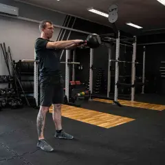 Mężczyzna ćwiczy z kettlebell w siłowni. W tle stojaki z ciężarami i sprzęt do treningu siłowego.