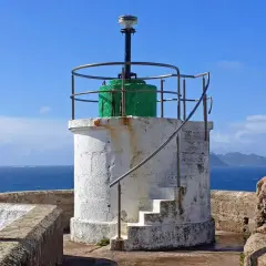 Faro blanco con base verde y escaleras externas, junto al mar azul. El **monte Ferro** se divisa al fondo.
