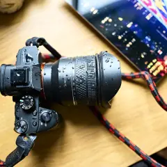 A Rokinon 24-28mm FE lens on a camera, covered in water droplets. The camera has a red and black strap. A tablet with a city lights display is in the background.